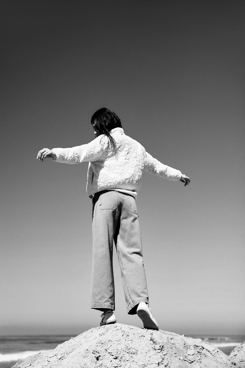 Person standing on a beach with arms outstretched, wearing a textured sweater and light-colored pants.